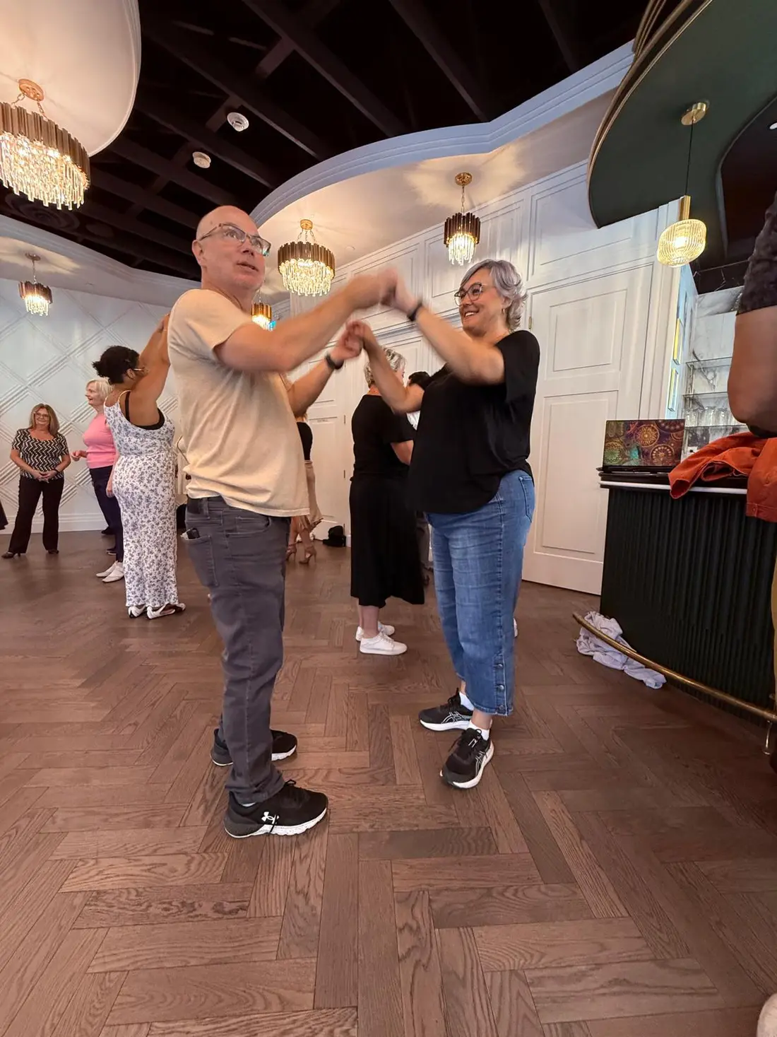 An older couple dancing hand in hand, both smiling, under the chandeliers